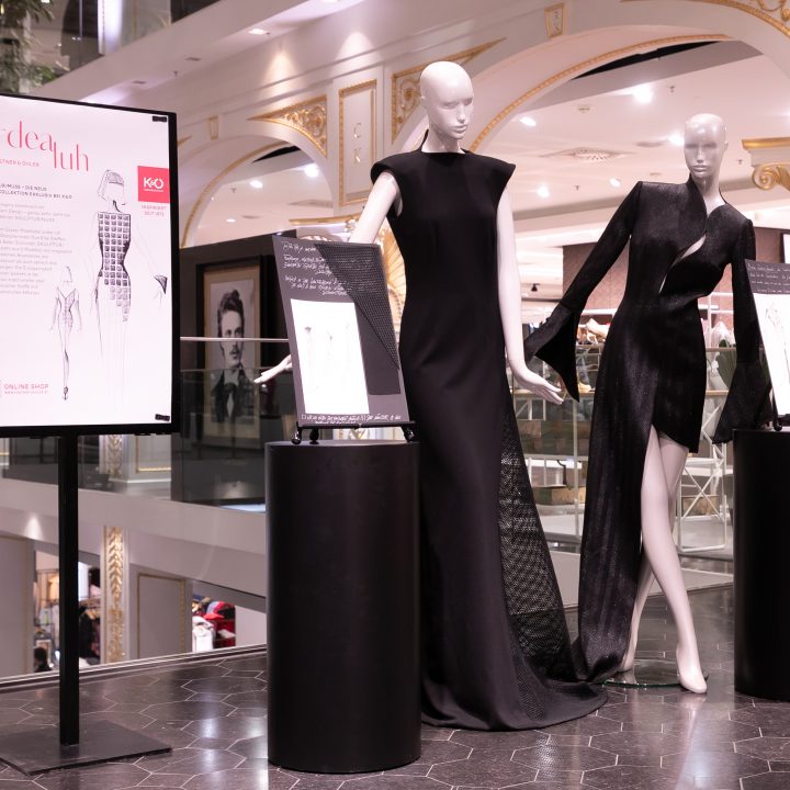 Dans une boutique de luxe, deux mannequins sont exposés dans d'élégantes robes noires. La mise en scène comprend un décor artistique et des panneaux d'information sur les tenues.