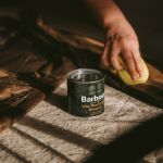 Une personne applique de la cire sur un vêtement marron avec une éponge jaune. Une boîte portant l'inscription « Barbour Wax Thornproof Dressing » se trouve à proximité sur une surface recouverte de tissu. La scène est éclairée chaleureusement, et les ombres suggèrent une source de lumière naturelle.