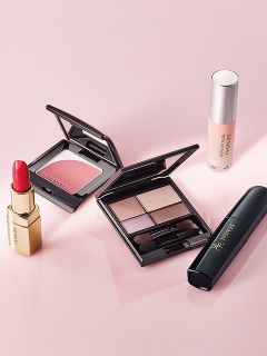 Une série de produits de maquillage, dont du rouge à lèvres, du fard à joues, une palette d'ombres à paupières, du mascara et du fond de teint, disposés sur une surface rose.