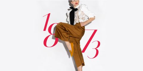 Une personne pose devant un fond blanc. Elle porte un haut en dentelle blanche, un foulard noir et un pantalon marron à jambes larges. Les chiffres rouges « 1873 » sont artistiquement superposés à l'image. Le visage de la personne est tourné sur le côté et n'est pas visible.