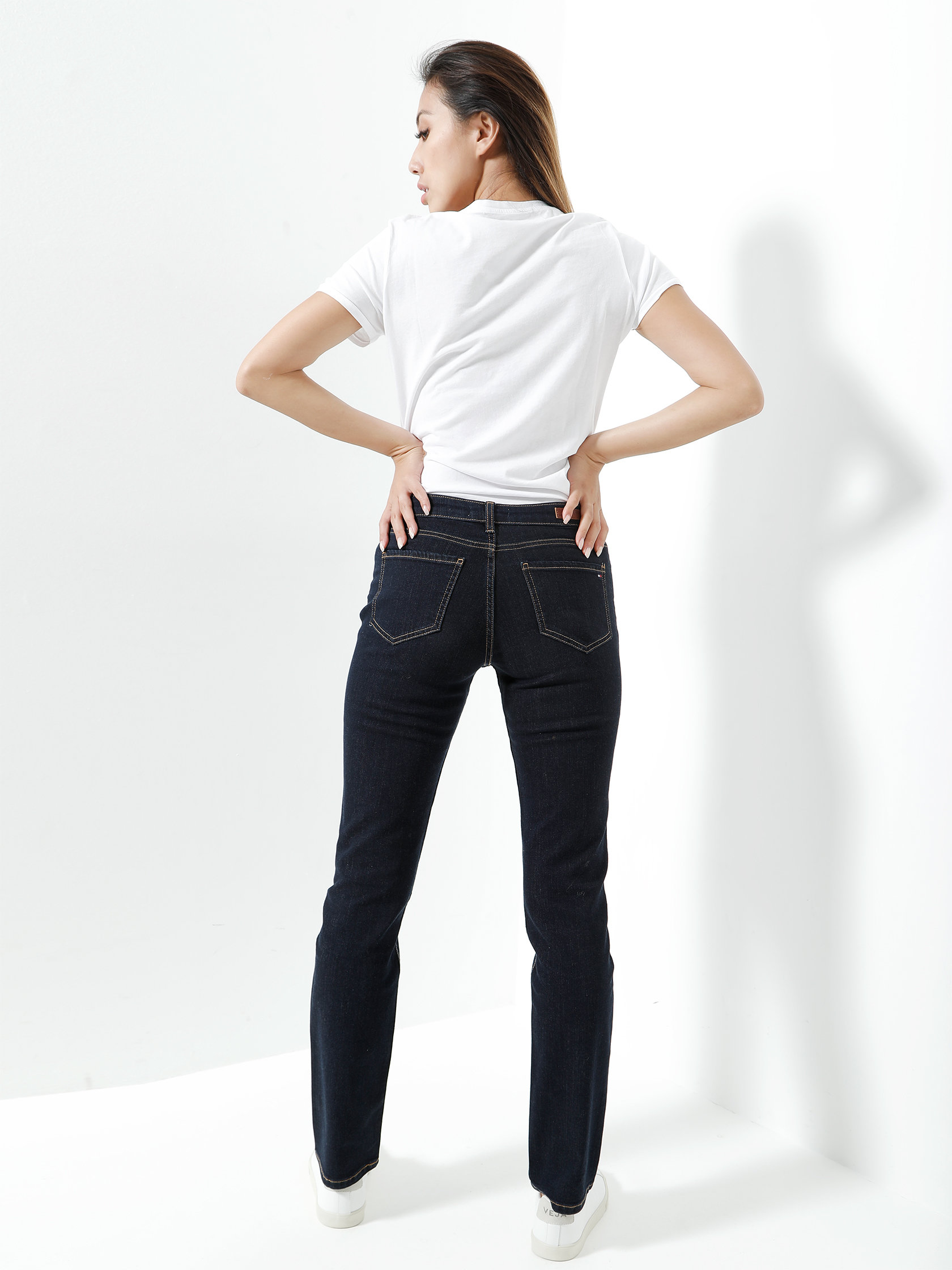 Une personne se tient devant un fond blanc et regarde au loin de la caméra. Elle porte un t-shirt blanc, un jean foncé et des baskets blanches, et a les mains sur les hanches.