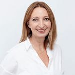Une femme aux cheveux châtain clair mi-longs sourit à la caméra. Elle porte un chemisier blanc et un collier doré et se tient devant un fond clair et uni.