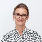 Une personne portant des lunettes sourit à la caméra. Elle porte une chemise blanche à pois noirs. L'arrière-plan est simplement blanc.