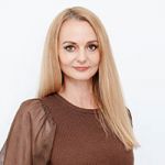 Une femme aux longs cheveux blonds et portant un haut marron se tient devant un fond blanc. Elle sourit légèrement et regarde directement la caméra.