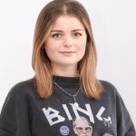 Une femme aux cheveux bruns mi-longs et portant un sweat-shirt noir à motifs graphiques se tient devant un fond blanc uni. Elle a une expression neutre et porte un collier argenté.
