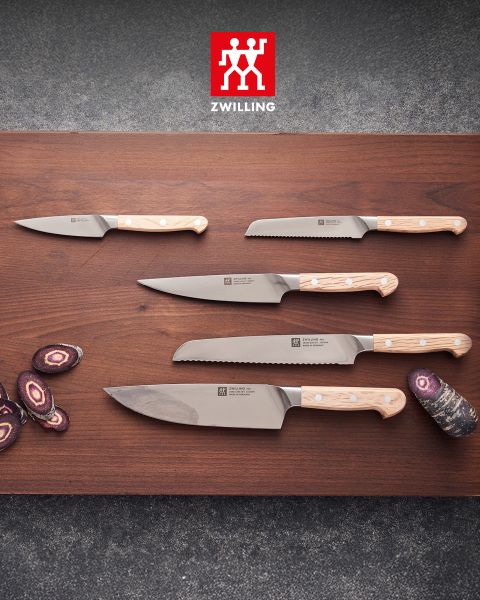 Cinq couteaux de cuisine avec des manches en bois clair sont posés sur un support en bois, à côté se trouvent des carottes violettes coupées en rondelles, et au-dessus, on peut voir le logo Zwilling.