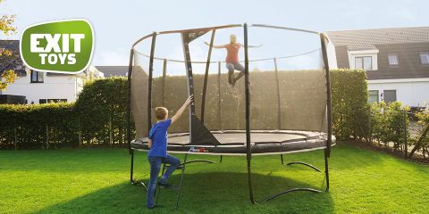 Deux enfants jouent sur un trampoline rond avec un filet de sécurité dans une cour arrière. Un enfant saute pendant que l'autre grimpe à une petite échelle. La scène se déroule par une journée ensoleillée avec des maisons en arrière-plan. Dans un coin, on peut voir le logo de « Exit Toys ».
