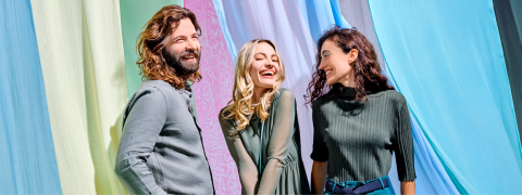 Trois personnes souriantes se tiennent devant des fonds en tissu colorés. L'arrière-plan est dans différentes nuances de vert, rose, violet et bleu. Tous portent des vêtements décontractés, ont l'air joyeux et discutent.