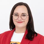 Une personne souriante aux longs cheveux foncés et portant des lunettes, vêtue d'un blazer rouge sur une chemise blanche, se tient devant un fond blanc uni.