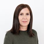 Une femme aux cheveux bruns mi-longs et portant un pull vert foncé se tient devant un fond blanc uni. Elle regarde l'appareil photo avec une expression neutre.