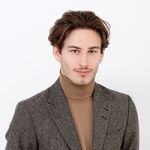 Une personne aux cheveux bruns et à la moustache porte un blazer gris sur un pull à col roulé beige. Elle sourit légèrement et regarde la caméra devant un fond blanc uni.