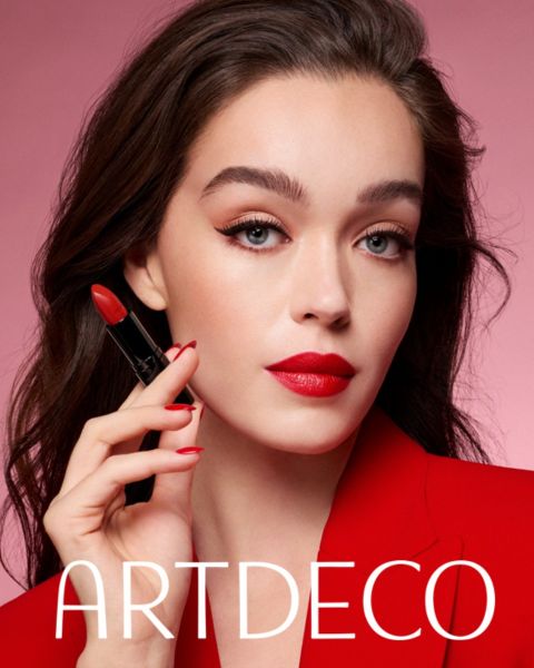 Une femme à la peau claire et aux cheveux foncés pose devant un fond rose, tenant un rouge à lèvres rouge et portant un blazer rouge ainsi qu'un rouge à lèvres rouge assorti. En dessous figure l'inscription « ARTDECO ».