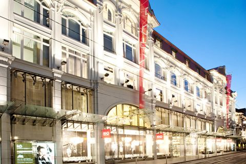 Façade illuminée d'un grand centre commercial élégant le soir, avec des fenêtres cintrées et des toits en verre. La rue est vide et les vitrines sont brillamment éclairées.
