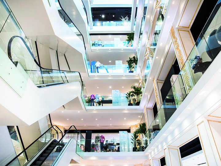 Vue intérieure d'un centre commercial moderne à plusieurs étages avec un éclairage vif, des balustrades en verre, des escaliers mécaniques et des espaces verts à chaque étage. La conception ouverte présente plusieurs étages et façades de magasins dans un environnement spacieux.