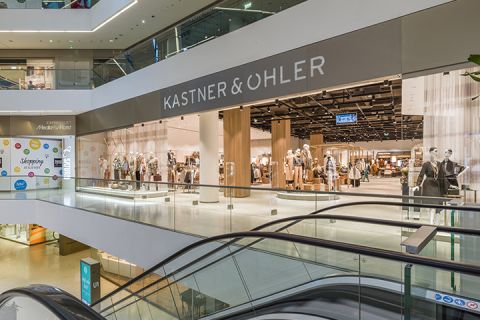 Vue intérieure d'un centre commercial moderne avec le magasin Kastner & Öhler. Des mannequins portant différentes tenues sont exposés sur la façade du magasin. Au premier plan, on aperçoit des escaliers mécaniques et le centre commercial comporte plusieurs niveaux avec des balustrades en verre.