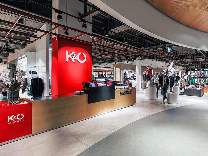 Vue intérieure d'un magasin de vêtements moderne avec un comptoir rouge vif portant le logo K&Ö. Plusieurs portants à vêtements sont visibles dans le magasin, avec des mannequins en arrière-plan et une personne qui regarde les articles. L'atmosphère est spacieuse et bien éclairée.