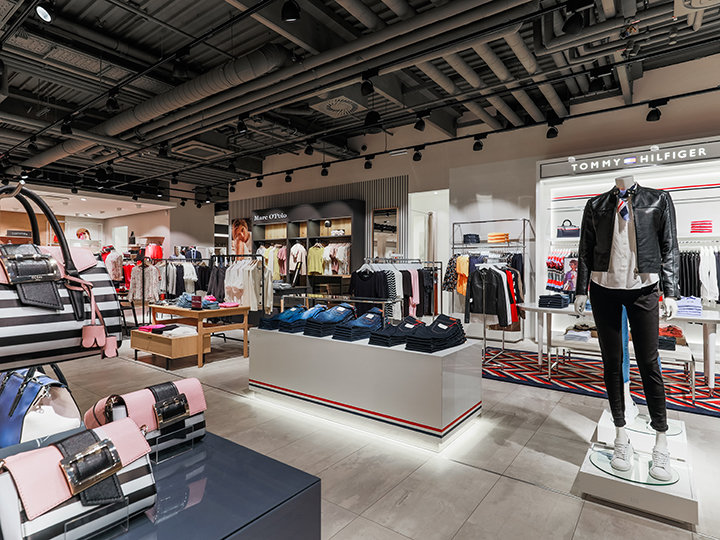 Un intérieur moderne d'un magasin de vêtements présentant divers articles d'habillement. Au premier plan, des mannequins présentent des tenues à la mode. Des vêtements pliés sont disposés sur des étagères et des accessoires sont exposés sur des tables. Un éclairage vif met en valeur le design moderne du magasin.