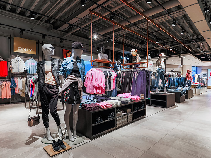 Vue intérieure d'un magasin de vêtements avec des mannequins portant des tenues décontractées, notamment des vestes et des jupes. Divers vêtements tels que des chemises et des jeans sont exposés sur des étagères et des présentoirs. Le magasin a un design moderne avec un éclairage au plafond et un sol carrelé.