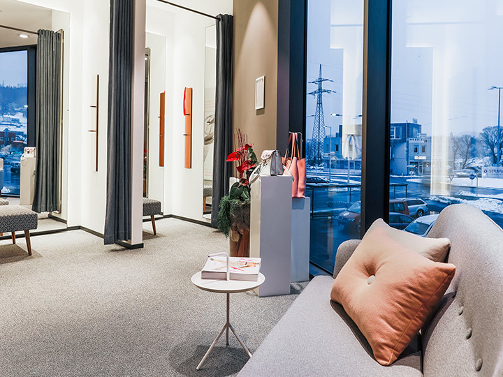 Une salle d'attente moderne et élégante avec un canapé, une petite table ronde et de grandes fenêtres offrant une vue sur un paysage urbain enneigé. La pièce est équipée de moquette, de rideaux et de plantes décoratives sur un socle.