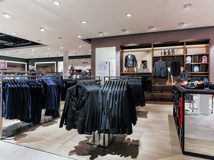 Une boutique de vêtements moderne avec des portants remplis de costumes soigneusement suspendus. Sur les étagères à l'arrière-plan, on peut voir des chemises pliées, des blazers et des accessoires. La boutique est bien éclairée et présente un agencement élégant et épuré.