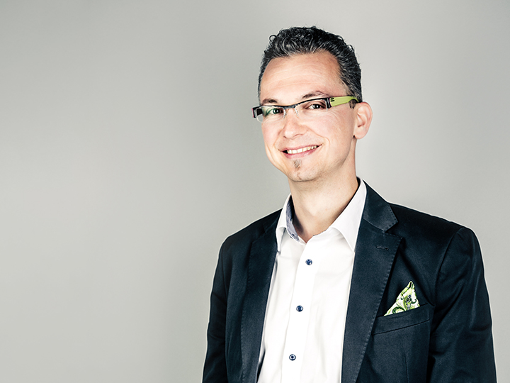 Un homme aux cheveux courts et foncés et portant des lunettes sourit à la caméra. Il porte un blazer foncé sur une chemise blanche avec une pochette de costume à motifs. L'arrière-plan est simplement gris clair.