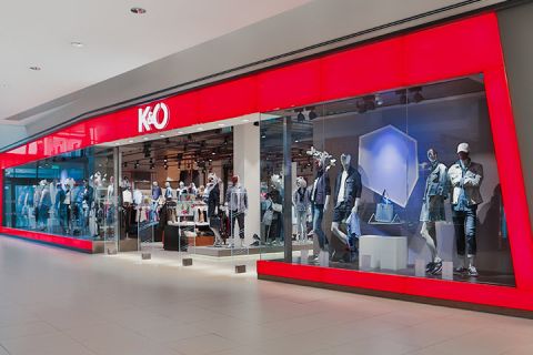 Vitrine avec une grande façade en verre rouge, où des mannequins sont exposés en tenues décontractées. L'intérieur est bien visible et montre des portants à vêtements. Le logo du magasin présente un « K&Ö » stylisé.