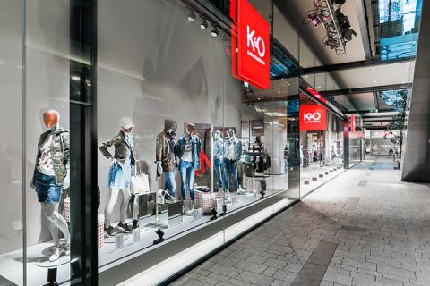Une vitrine d'un magasin de mode présentant des mannequins portant des tenues décontractées et tendance, notamment des vestes et des shorts en jean. Le magasin a de grandes vitrines et est situé dans un centre commercial moderne à l'architecture élégante.