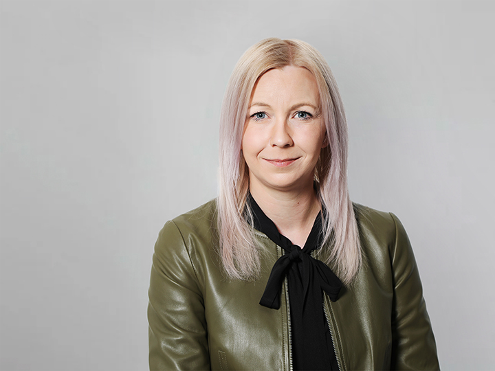 Portrait d'une femme aux cheveux lisses et blond clair, portant une veste en cuir verte sur un haut noir. Elle sourit subtilement devant un fond uni gris clair.