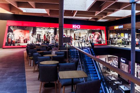 L'intérieur d'un centre commercial moderne avec un magasin de vêtements éclairé en rouge appelé K&O. L'espace comprend des coins salons avec des chaises et des tables, un escalator et diverses autres vitrines en arrière-plan.
