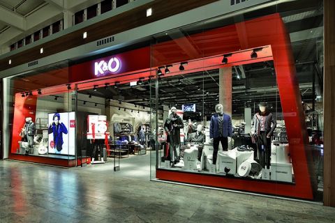 Vitrine de K&Ö dans un centre commercial avec des mannequins portant différents styles vestimentaires. L'exposition comprend des vestes, des chemises et des accessoires sous un éclairage vif, d'autres vêtements sont visibles à l'intérieur.