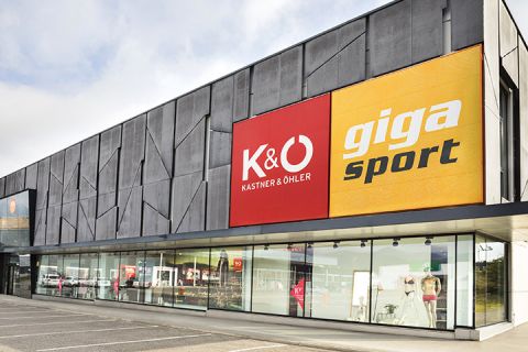 Vue extérieure d'un bâtiment moderne avec de grandes enseignes "Kastner & Öhler" en rouge et "Giga Sport" en jaune. Au rez-de-chaussée, de grandes vitrines présentent des articles de sport et de mode. Le ciel est couvert.
