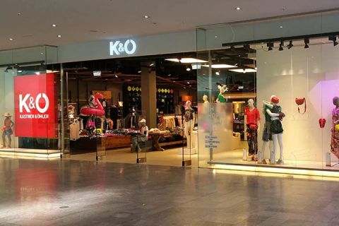 Vitrine de Kastner & Öhler avec de grandes vitres montrant des mannequins habillés. À l'intérieur du magasin, on peut voir des portants à vêtements et une sélection de vêtements. Le logo K&O brille bien visible au-dessus de l'entrée.