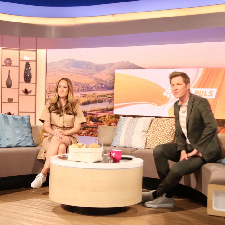 Un homme et une femme sont assis devant une télévision moderne avec un canapé en forme de U et des coussins décoratifs. Une boîte et une tasse sont posées sur une table basse. En arrière-plan, on aperçoit un paysage pittoresque. Des projecteurs de studio sont visibles au-dessus.