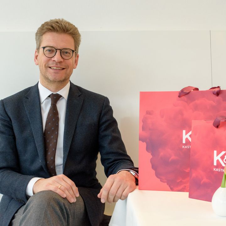 Un homme en costume et lunettes est assis à une table, souriant et tenant un sac de courses rouge. Il y a aussi un petit vase en céramique blanche sur la table.