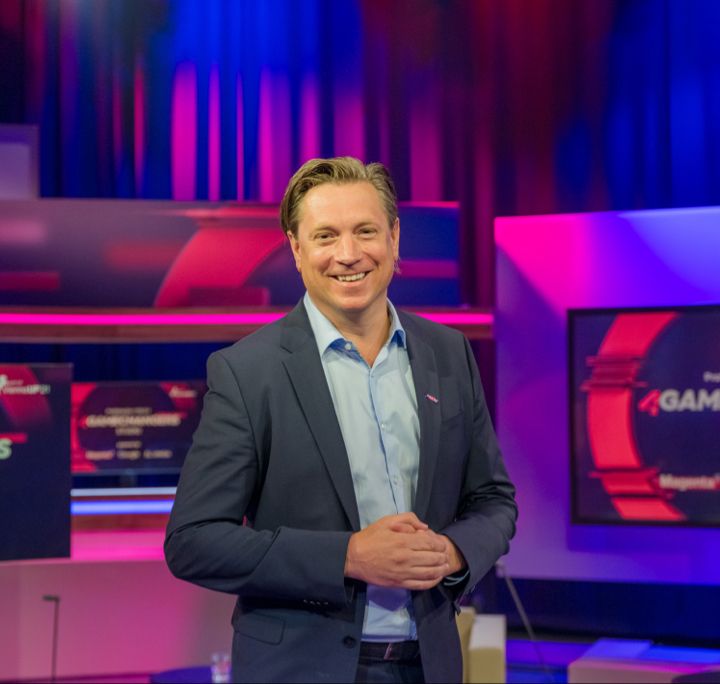 Un homme en costume se tient souriant dans un studio bien éclairé. Derrière lui, des écrans affichent le texte « GAMECHANGERS STUDIO » et les logos de Google et d'une autre entreprise. L'environnement est caractérisé par des lumières vives et colorées et une atmosphère professionnelle y règne.