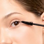 Eine Nahaufnahme einer Person, die schwarze Wimperntusche auf ihre oberen Wimpern aufträgt. Der Hintergrund hat eine neutrale Farbe und der Fokus liegt auf dem Auge und der Wimperntuschebürste.