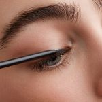 Nahaufnahme einer Person, die schwarzen Eyeliner auf ihr oberes Augenlid aufträgt. Das Auge ist teilweise geöffnet und zeigt gepflegte Augenbrauen und lange Wimpern. Der Eyeliner-Stift ist für Präzision angewinkelt.