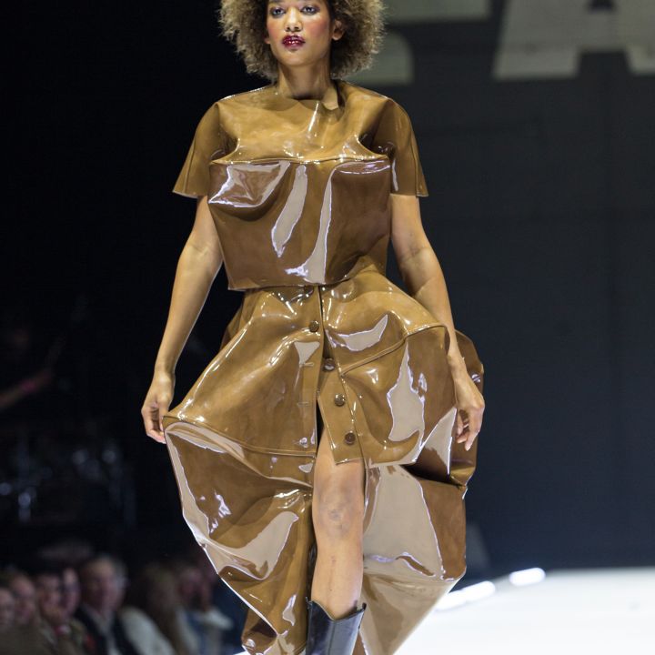 Un mannequin défile sur le podium, portant un haut brillant brun, comme en plastique, et une jupe, avec des bottes noires. L'arrière-plan est sombre et l'on aperçoit partiellement un public qui assiste au défilé de mode.