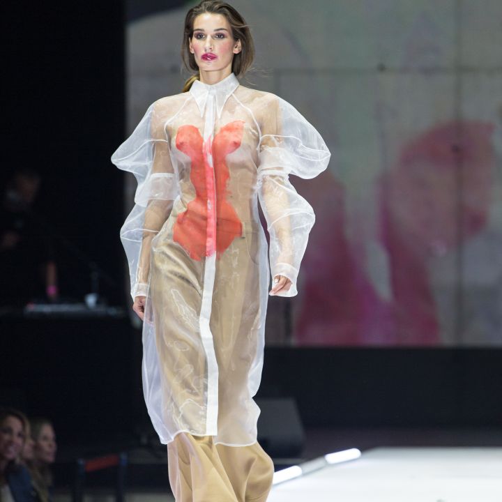 Un mannequin défile sur un podium, portant une robe blanche transparente à manches bouffantes et intérieur orange, ainsi qu'un pantalon beige. En arrière-plan, une image floue est visible, et des spectateurs sont visibles sur les côtés.