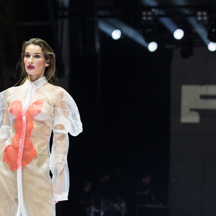 Un mannequin défile sur le podium, portant une robe transparente à manches longues avec de fortes touches de rouge, sous une lumière de scène vive. L'arrière-plan est une toile de fond sombre avec un grand panneau partiellement visible.