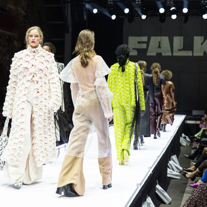 Des mannequins défilent sur un podium lumineux dans différentes tenues à la mode, notamment un ensemble floral et un vêtement transparent. Un public nombreux regarde, avec "FALK" affiché en arrière-plan.
