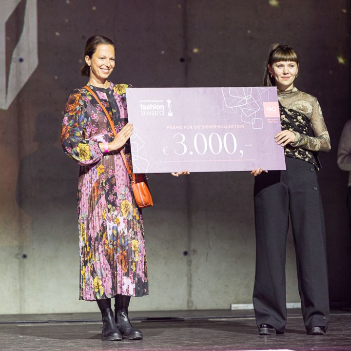 Deux femmes se tiennent sur scène lors d'une remise de prix de la mode et tiennent un grand chèque rose de 3 000 euros. L'une porte une robe à fleurs, l'autre un pantalon noir et un haut. Un logo K&O est visible en arrière-plan, une autre personne se tient plus loin derrière.