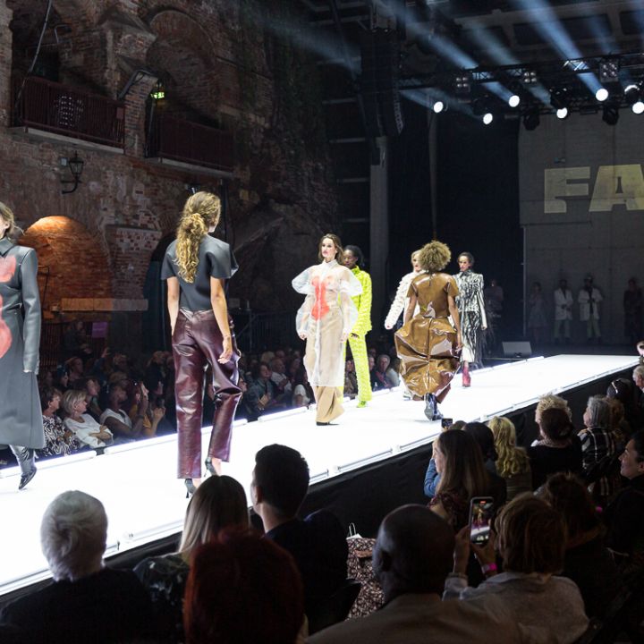 Lors d'un défilé de mode, des mannequins défilent sur le podium, présentant des créations colorées et artistiques. La salle est faiblement éclairée, avec des spectateurs assis de part et d'autre. Un grand panneau portant l'inscription "FALK" brille en arrière-plan.