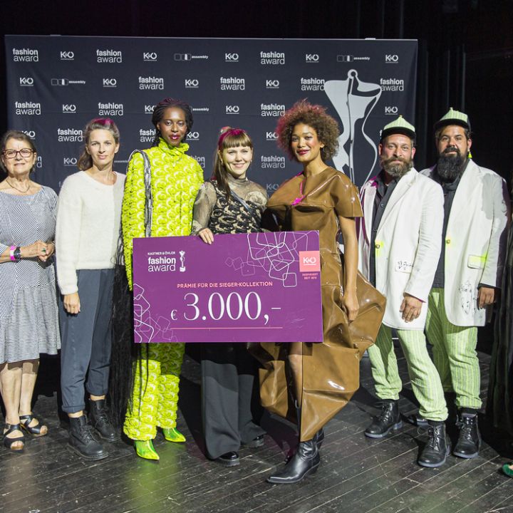 Un groupe de dix personnes se tient sur une scène, dont une personne tenant un grand chèque de prix violet de 3 000 dollars. Ils portent des tenues chics et colorées. Derrière eux, on peut voir une toile de fond avec le nom de l'événement.