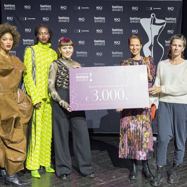 Cinq personnes se tiennent sur une scène et tiennent un grand chèque de 3 000 euros. En arrière-plan, on aperçoit le logo d'un prix de la mode. Elles sont vêtues de tenues élégantes et colorées et semblent faire la fête.