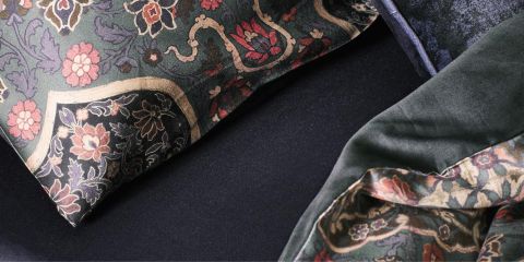 Gros plan d'un lit avec des coussins foncés à motifs floraux et une couverture à motifs assortis. Le tissu présente des motifs complexes dans des tons de rouge, rose et vert, qui ressortent sur un fond sombre. Le tissu a l'air moelleux et doux.