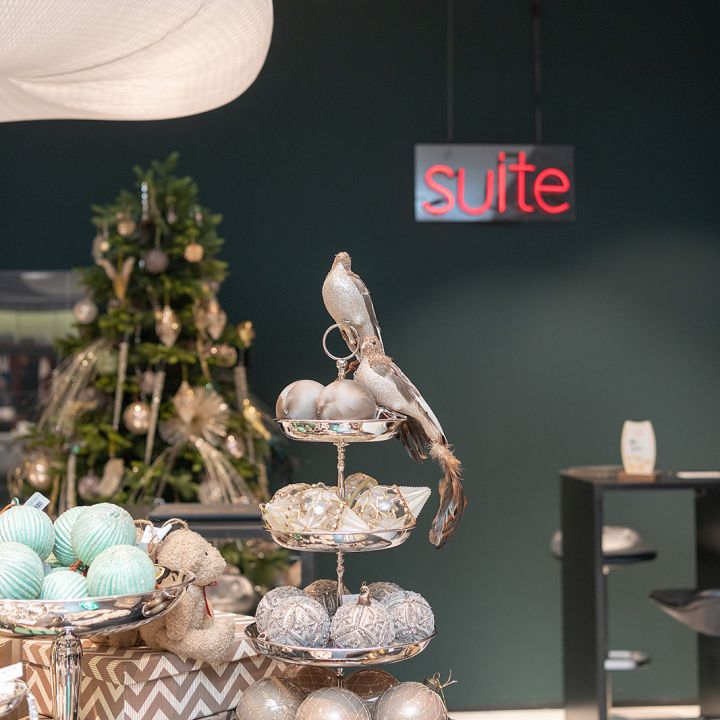 Une présentation festive comprend un plateau à plusieurs étages avec des boules décoratives et un ornement d'oiseau. En arrière-plan, un sapin de Noël est décoré de lumières et d'ornements. Un néon rouge avec l'inscription « Suite » est accroché à un mur vert foncé.