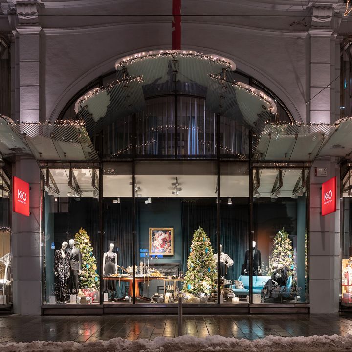 Une vitrine festive avec des décorations de Noël, notamment des lumières scintillantes, des sapins de Noël et des mannequins portant des vêtements d'hiver. La fenêtre est encadrée par une structure arquée et le sol à l'extérieur est légèrement recouvert de neige.