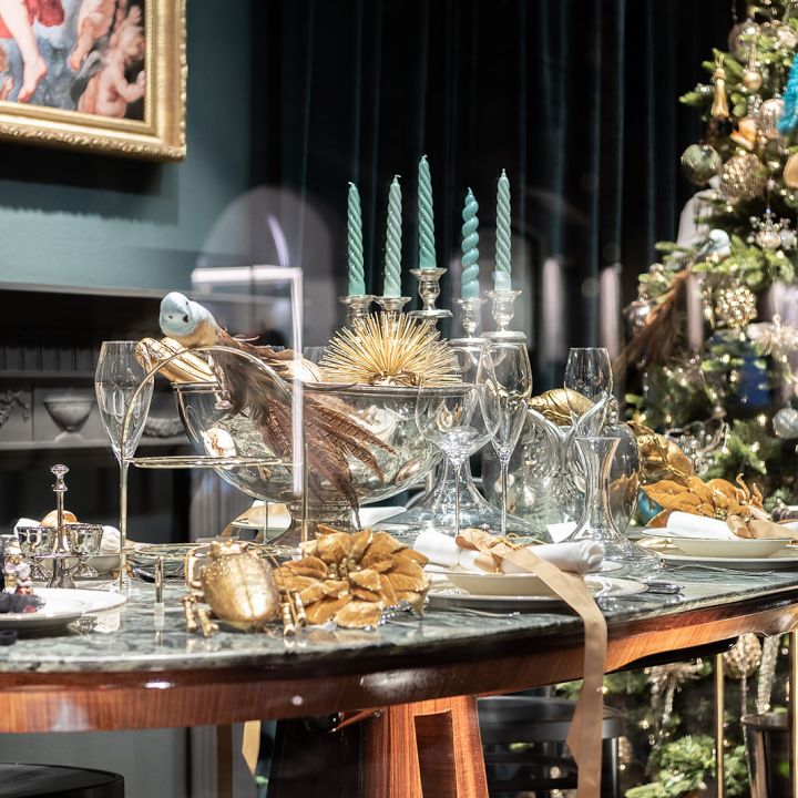 Une table à manger richement décorée pour une occasion festive avec des bougies vertes, des accents dorés, des verres en cristal et un luxueux sapin de Noël en arrière-plan. L'ambiance est élégante avec un mur sombre et un tableau encadré.
