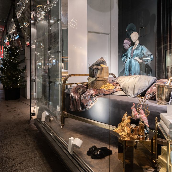 Une vitrine présentant un lit élégamment décoré et divers articles de luxe tels que des sacs à main et un seau à champagne. Des illuminations et décorations de Noël ornent la rue à l'extérieur, créant une ambiance festive.